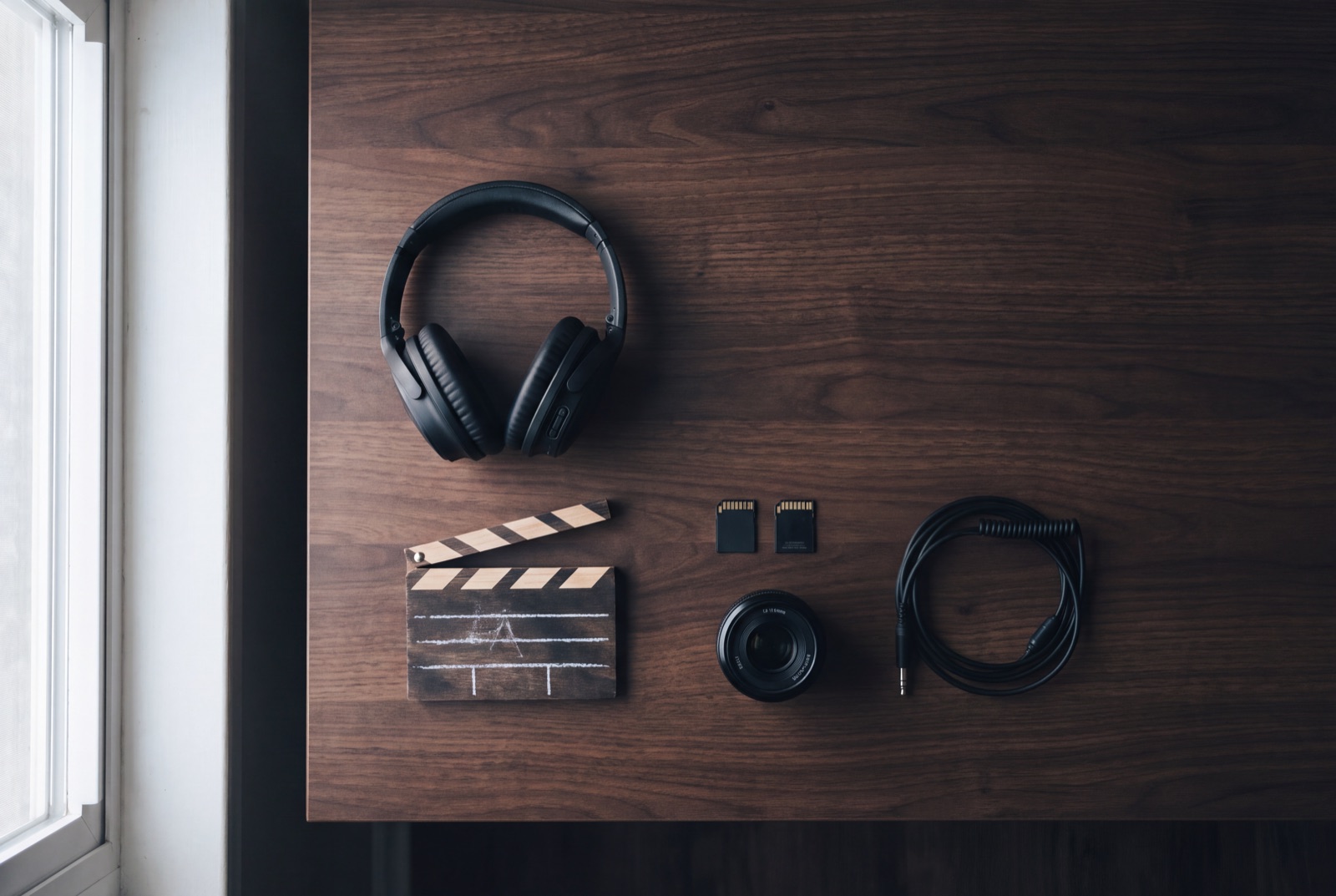 A flat lay of headphones, a clapperboard, SD cards and a lens on a dark desk